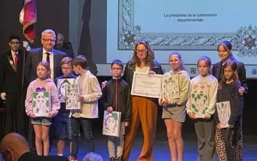 enfants tenant leurs réalisation, enseignantes tenant le diplôme, membres de l'association et porte-drapeaux sur la scène avec diplôme projeté sur le mur à l'arrière. 